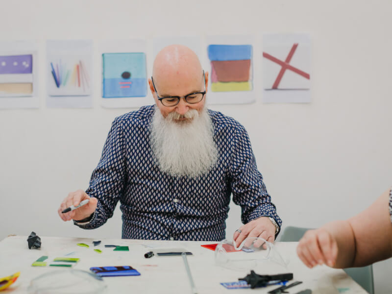 Man with large white beard measuring out glass pieces with a sharpie and ruler for a mosaic.